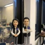 兩尊成對的人像公仔置於透明展示盒外，由手掌襯托拍攝：皆穿白色襯衫、深色無袖背心與淺灰長褲、白色運動鞋。左側為長髮女性公仔，雙手在腹前交握、微笑；右側為短髮男性公仔，右臂環抱左臂呈自信站姿。兩者胸前皆別有深藍識別證牌；腰間可見皮帶與簡化裝備造型。公仔站在石磚紋方形台座，前方金色名牌各以黑字刻有稱呼與致意語。背景是室內工作室環境與窗邊夜色反射，營造致敬與送禮紀念氛圍。