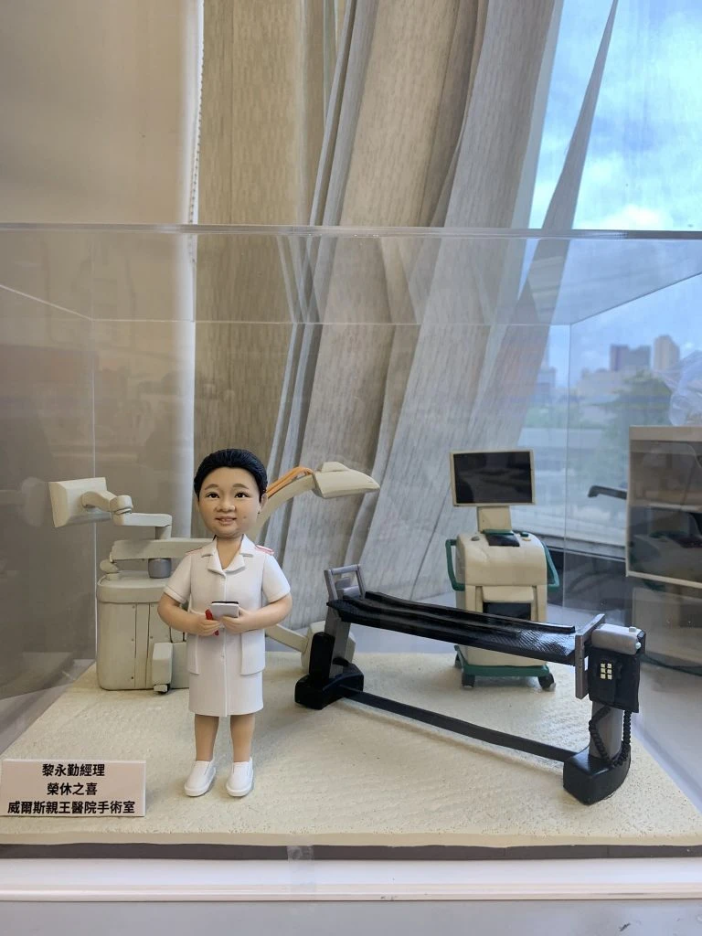 手工訂製醫護人像公仔，人物身穿白色醫護制服，站立於精緻的手術室場景模型中。背景包括手術燈、醫療儀器及手術檯，細節逼真。底座文字顯示：「黎永勤經理 經休之喜 威爾斯親王醫院手術室」，象徵專屬醫生與醫護人員的定製禮物，用於紀念醫療團隊的專業與感謝之情。