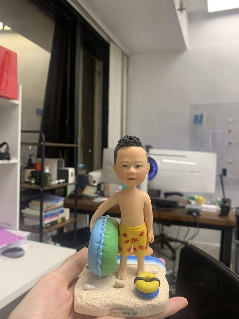 手掌上托著一尊兒童造型的游水人像公仔。公仔留短髮，赤裸上身，穿著黃色印有紅色小魚圖案的泳褲，右手自然下垂，左臂抱著綠藍雙色圓形泳圈。腳踩貝殼與細沙質感的底座，旁邊放著藍黃配色的小蛙鞋與小水池邊緣造型。Keywords：人像公仔，游水人像公仔。