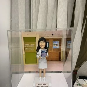手工訂製醫護人像公仔，人物穿著白色醫護制服，手持文件夾與毛公仔，底座寫有「Annie Leung Happy Retirement」。背景是醫院內的「綜合日間服務 / 內分泌及糖尿科日間護理部」門口，顯示此公仔專為糖尿科醫生或醫護人員榮休而製作，是獨特且具紀念價值的專屬禮物。
