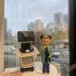 醫生人像公仔，身穿綠色手術袍與藍色醫師服，戴著眼鏡微笑站立，手持內視鏡操作器械。旁邊有超聲波內視鏡診斷儀器與螢幕，場景高度還原臨床醫療環境。此人像公仔是專屬醫生、中醫師的客製化禮物，適合送給醫護人員作為紀念或感謝之用。
