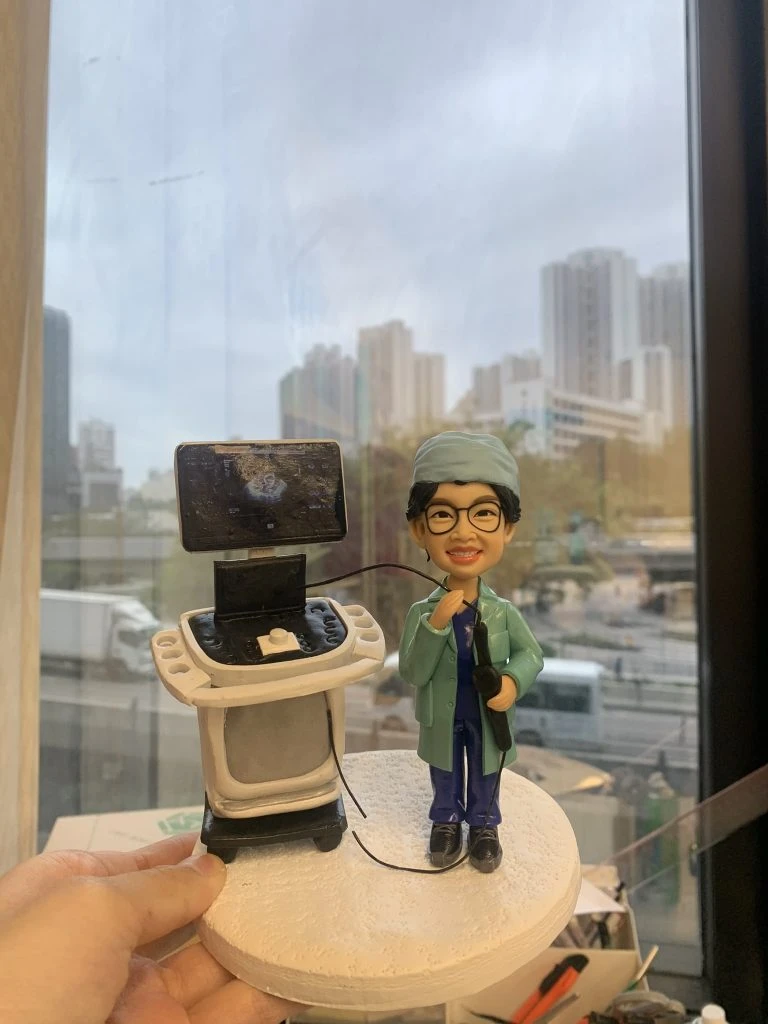 醫生人像公仔，身穿綠色手術袍與藍色醫師服，戴著眼鏡微笑站立，手持內視鏡操作器械。旁邊有超聲波內視鏡診斷儀器與螢幕，場景高度還原臨床醫療環境。此人像公仔是專屬醫生、中醫師的客製化禮物，適合送給醫護人員作為紀念或感謝之用。