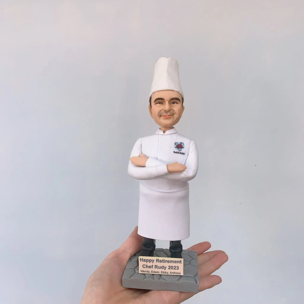 手掌托起的廚師主題人像公仔：戴高帽、穿全白廚師服的男廚師雙臂交叉微笑站立，胸口左側繡有彩色圖案與姓名字樣。腳下是灰色石磚紋底座，前方金色名牌刻著「Happy Retirement Chef Rudy 2023」及贈送者名字。背景為素色牆面，焦點集中在公仔細節與典禮感十足的退休題字，呈現典雅的廚師退休禮物擺設。