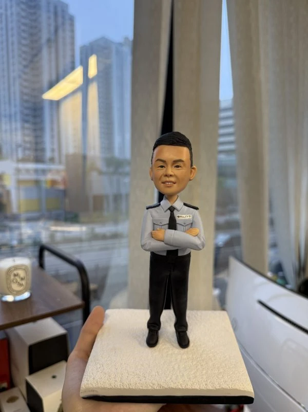 一尊以「督察」職級為主題的警察人像公仔，被手掌托於室內窗邊。公仔短黑髮、微笑神情；身穿淺藍色長袖制服襯衫，配黑色領帶與深色肩章，左胸名牌旁印有「POLICE」字樣；雙臂於胸前交叉。下身為黑色長褲與黑色皮鞋。人物站在淺米色粗糙質感的方形底座上。背景為落地窗外的城市街景：密集高樓、道路與行道樹；玻璃上可見長條燈的反射。室內有米色窗簾、木質小桌與蠟燭杯，以及白色設備邊角。整體呈現在辦公室窗邊展示、便於送禮與陳列的情境。