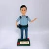 一尊以「警員」職級為主題的人像公仔置於白色無縫背景前拍攝。公仔黑色短髮、微笑神情；身穿淺藍色短袖制服襯衫，肩上有深色肩墊與扣件，左胸口袋上方印有 POLICE 字樣。腰間繫黑色裝備腰帶，配有多個裝備袋與扣具；下身為深藍長褲與黑色皮鞋。右手自然下垂並持一頂深藍色頭盔，左臂抬起向側前方打開手掌做示意/招呼姿態。腳下為深綠色方形台座，前側裝有米金色名牌，刻有「爸爸退休快樂」等祝福字樣，台座兩角點綴紅色小花。整體以簡潔白底呈現，凸顯公仔與制服細節，營造紀念與致意的送禮場景。