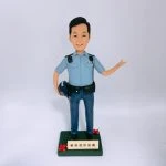 一尊以「警員」職級為主題的人像公仔置於白色無縫背景前拍攝。公仔黑色短髮、微笑神情；身穿淺藍色短袖制服襯衫，肩上有深色肩墊與扣件，左胸口袋上方印有 POLICE 字樣。腰間繫黑色裝備腰帶，配有多個裝備袋與扣具；下身為深藍長褲與黑色皮鞋。右手自然下垂並持一頂深藍色頭盔，左臂抬起向側前方打開手掌做示意/招呼姿態。腳下為深綠色方形台座，前側裝有米金色名牌，刻有「爸爸退休快樂」等祝福字樣，台座兩角點綴紅色小花。整體以簡潔白底呈現，凸顯公仔與制服細節，營造紀念與致意的送禮場景。