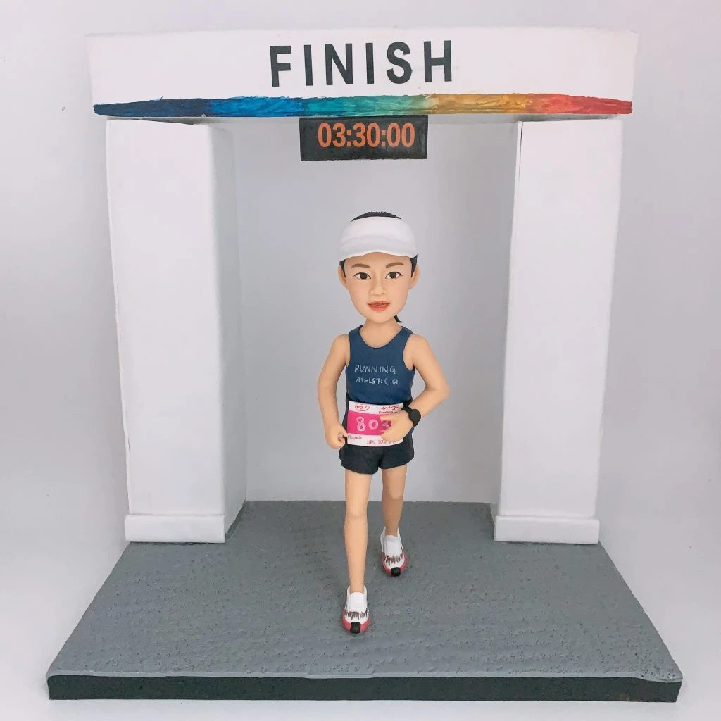 馬拉松比賽主題人像公仔：女跑者戴白色遮陽帽，穿深藍無袖跑衣與黑色短褲，胸前繫粉紅號碼布，腳踏白紅跑鞋；她正從寫有「FINISH」字樣的終點拱門下跑出，拱門下方電子計時牌顯示「03:30:00」，灰色顆粒地台模擬賽道質感。整體呈現完成馬拉松的高光時刻，適合作為馬拉松紀念與收藏的人像公仔。