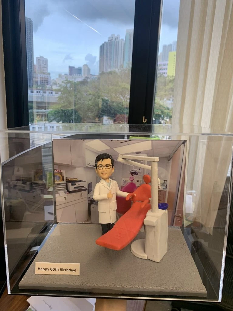 一座放在透明展示盒內的牙科主題人像公仔：公仔身穿白色醫生袍與眼鏡，右手比讚，站在紅色牙科治療椅旁；椅側配有牙科器械柱、口腔吸引系統與控制面板，頭頂延伸手臂式無影燈；背景為診療室櫥櫃與儀器照片。底座前方小牌寫有「Happy 60th Birthday!」，呈現為牙科醫護人員的客製紀念禮物。