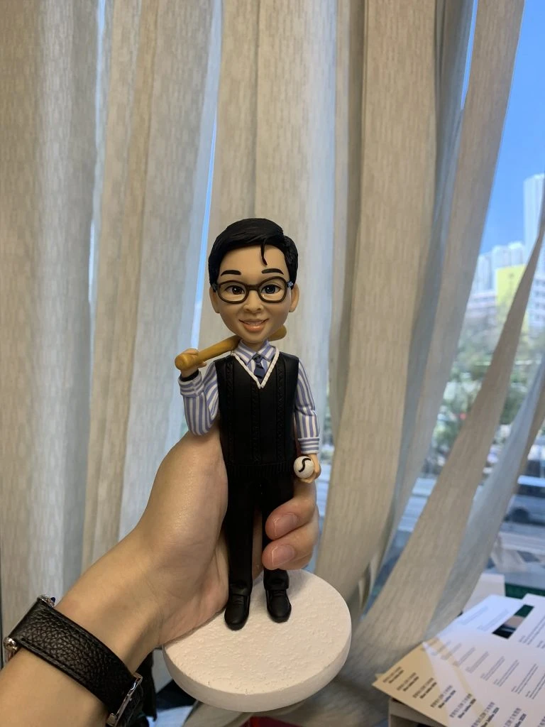 一款棒球主題的人像公仔：短黑髮、戴黑框眼鏡的男子露出微笑，穿著藍白直條襯衫、黑色針織背心與西裝長褲，左手握有棒球，右肩自然扛著木質球棒；腳踏黑色皮鞋，站在白色圓形底座上。背景為室內窗簾與城市景色的淺景深，前景可見手掌托起公仔。此作品強調職場紳士風與運動元素結合的棒球人像公仔與人像公仔。