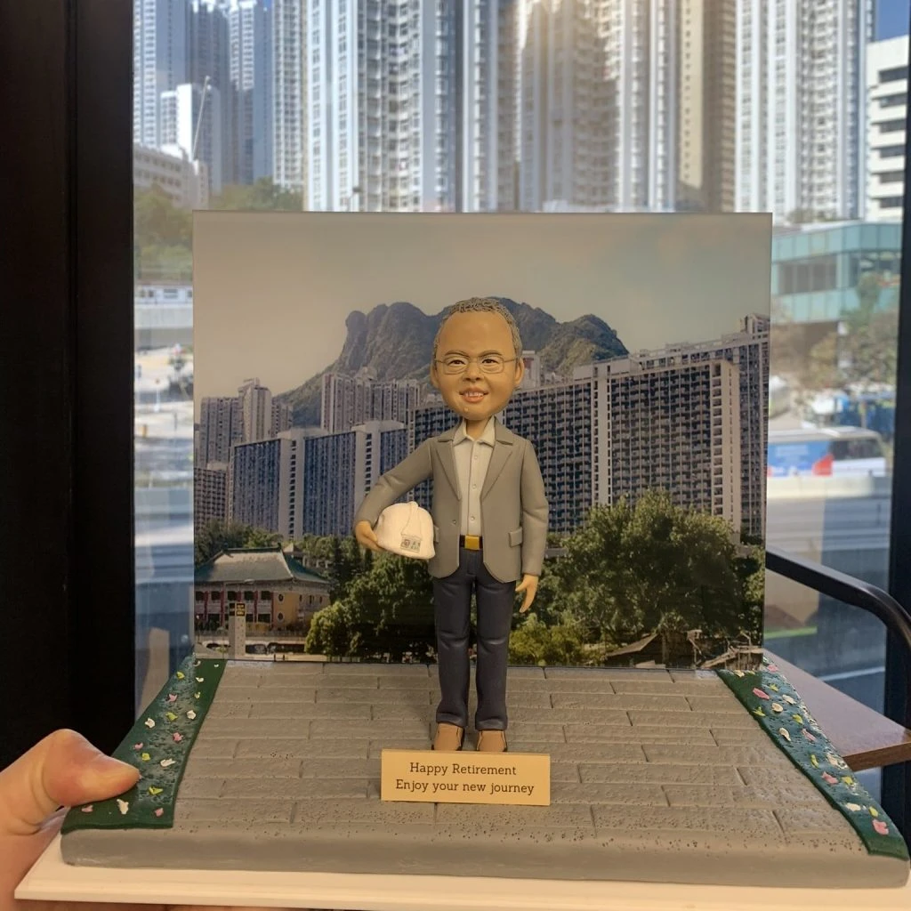 一個以香港獅子山為背景的榮休禮物。模型主角是一位戴眼鏡、身穿西裝、手持安全帽的人像公仔。他站在以獅子山和周邊住宅樓宇為背景的展板前,是一份向建設社區的工程師致敬的禮物,銘牌上寫有退休祝福。