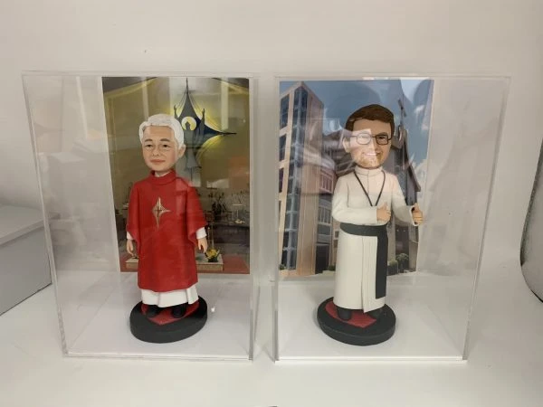 兩款並排展示在透明亞加力膠盒中的客製化神父人像公仔，左邊身穿紅色祭披，右邊身穿白色聖袍比讚，是送給牧師或神職人員的禮物系列。