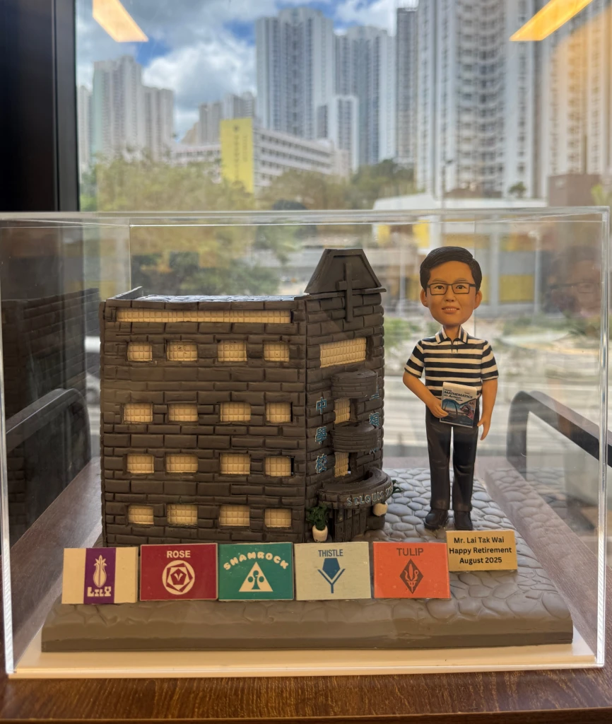 一款為聖類斯中學的黎德維校長（Mr. Lai Tak Wai）訂製的校長退休禮物。精緻的人像公仔手持數學書，莊重地站在一座細節豐富的聖類斯中學立體學校模型旁，底座前方陳列著五個社（LILY, ROSE, SHAMROCK, THISTLE, TULIP）的標誌，整件作品置於透明保護罩中，是一份極具份量的榮休紀念品。