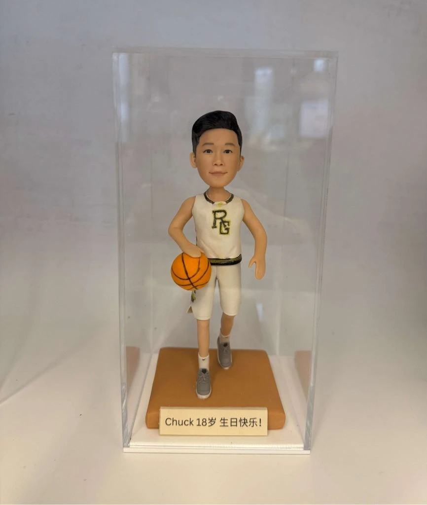 籃球員主題人像公仔：短髮少年公仔身穿白色籃球球衣與球褲，胸前有金邊字母標誌，腳踩灰色球鞋；右手持橙色籃球、左腳邁步向前的運球姿勢。公仔站在橘棕色方形底座上，前方銘牌寫有「Chuck 18岁 生日快乐！」字樣；整體置於透明展示盒中，白色背景簡潔，呈現適合作為生日禮物的籃球主題人像公仔。