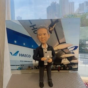航空主題的人像公仔退休禮物：灰白短髮男士公仔穿深色外套與淺灰襯衫，手抱平板電腦站在灰色質感底座上，前方名牌寫有「Congratulations on your 50 years of service / Chiu Hoi Wing」；背景板為停機坪與大型客機尾翼、機庫外牆與HAECO標誌，作品於室內靠窗拍攝，畫面下方可見手掌托住底座，體現向老闆與CEO致敬的榮退紀念。
