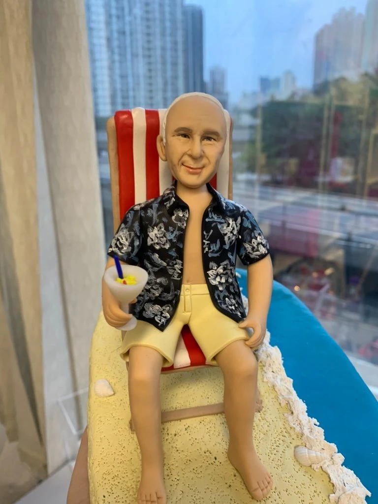 沙灘主題的人像公仔退休禮物:中年至長者男性公仔留短髮、面帶微笑,穿著黑底白花的夏威夷襯衫與米黃色短褲,赤腳坐在紅白條紋海灘椅上,右手握著帶藍色吸管與檸檬片的雞尾酒杯;腳邊細緻呈現米色沙灘、白色浪花邊與貝殼裝飾,旁有黃藍色救生圈小物。底座呈長形沙洲延伸至藍色海面,整體氛圍開心放鬆。拍攝於室內靠窗,背景可見城市高樓與道路景色,近距離與側拍角度皆強調度假感。