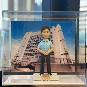 一款為香港女警員訂製的榮調紀念人像公仔，公仔身穿警察制服，背景為荔枝角收押所，由其同事為祝賀她調職而敬贈。