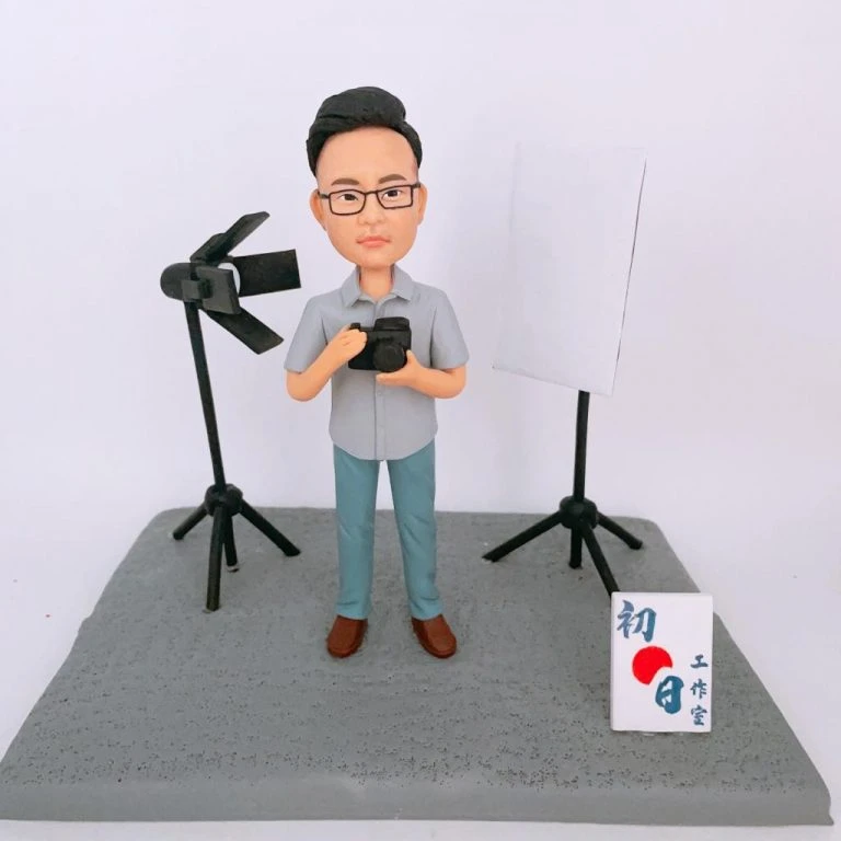 攝影記者主題人像公仔：短髮戴眼鏡的男士穿淺灰襯衫與藍綠長褲，雙手持相機站在灰色地台上；左側是帶遮光葉片的黑色攝影燈，右側為白色反光板與燈架，前方立有寫著「初日工作室」的白色小立牌。場景呈現採訪與拍攝現場氛圍，適合作為記者人像公仔榮休禮物與紀念擺設。