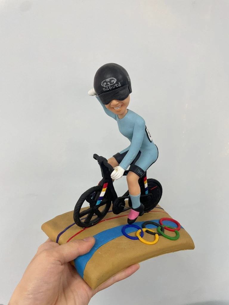 定制單車運動公仔，人物戴黑色安全帽與護目鏡，身穿天藍色緊身運動服，騎著黑色競賽自行車，底座為賽道設計並附有奧運五環彩色裝飾。
