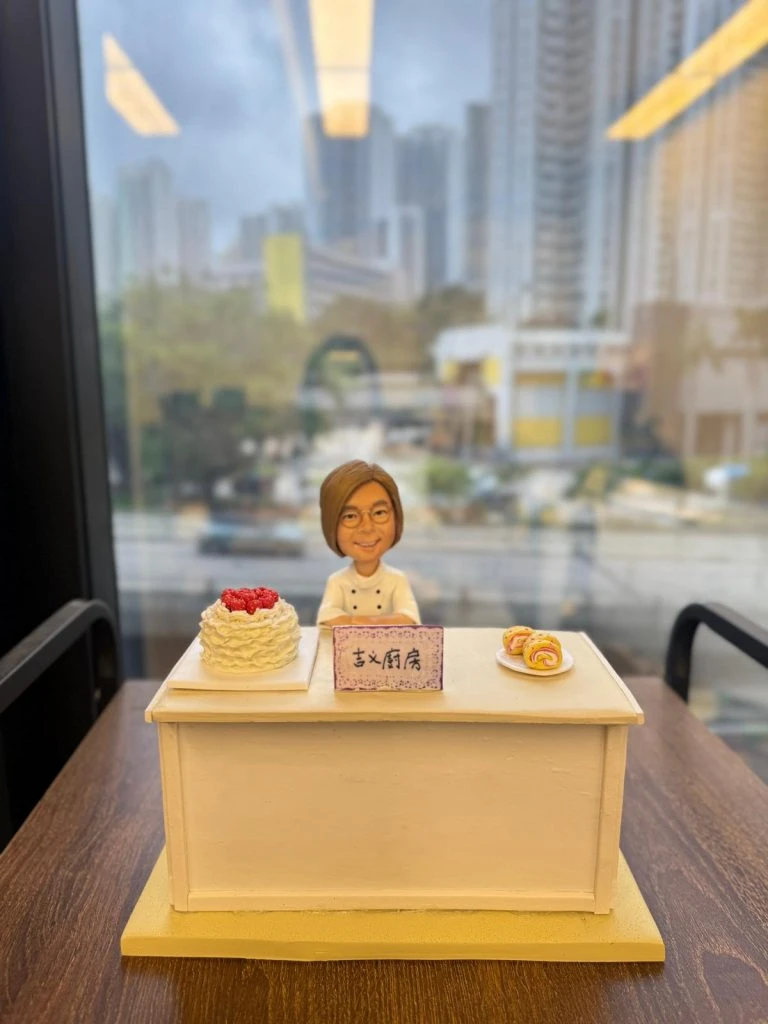 甜品店主題人像公仔擺設:短髮戴眼鏡的女主廚穿白色廚師服,站在淺米色櫃台後方微笑;台面左側放一個覆盆子奶油蛋糕,右側白盤擺放三枚捲蛋糕;正中央立有紫邊小立牌,手寫「玄X廚房」字樣(字跡帶手作感)。整體置於淺黃色底板與木紋桌面上,背後是窗景與高樓大廈的城市背景。造型溫馨且具儀式感,適合作為甜品開張禮物與店內展示。