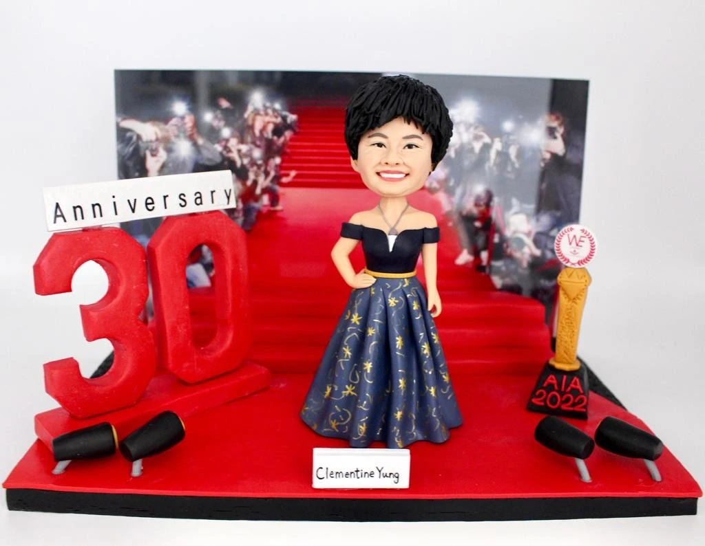 一座慶祝30周年的人像公仔場景：短髮女子微笑站立於紅毯階梯前，穿深藍色露肩禮服，裙擺以金色星月與圖騰點綴，腰間金色腰帶。左側有巨大紅色「30」立體字與「Anniversary」字牌，兩側設黑色欄杆/燈具；右側擺金色獎座，黑紅底座上寫「AIA 2022」。背景是紅毯上媒體攝影群的照片牆，前方白色名牌顯示姓名，整體營造隆重的里程碑慶典氛圍。