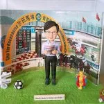 馬場人像公仔展示：戴眼鏡、穿淡紫襯衫與深色長褲的男子公仔雙手拿報紙站在綠色草地底座上；背板為沙田馬場場景，拱門寫有「Sha Tin Racecourse 沙田馬場」與賠率看板，右側可見賽馬奔馳畫面。前景擺有足球造型小物、寫著「Good Health」「Stay Happy」的路牌、金色獎座與小型花束裝飾，前方銘牌刻有感謝語。整體呈現賽馬與祝賀主題的馬場人像公仔收藏。