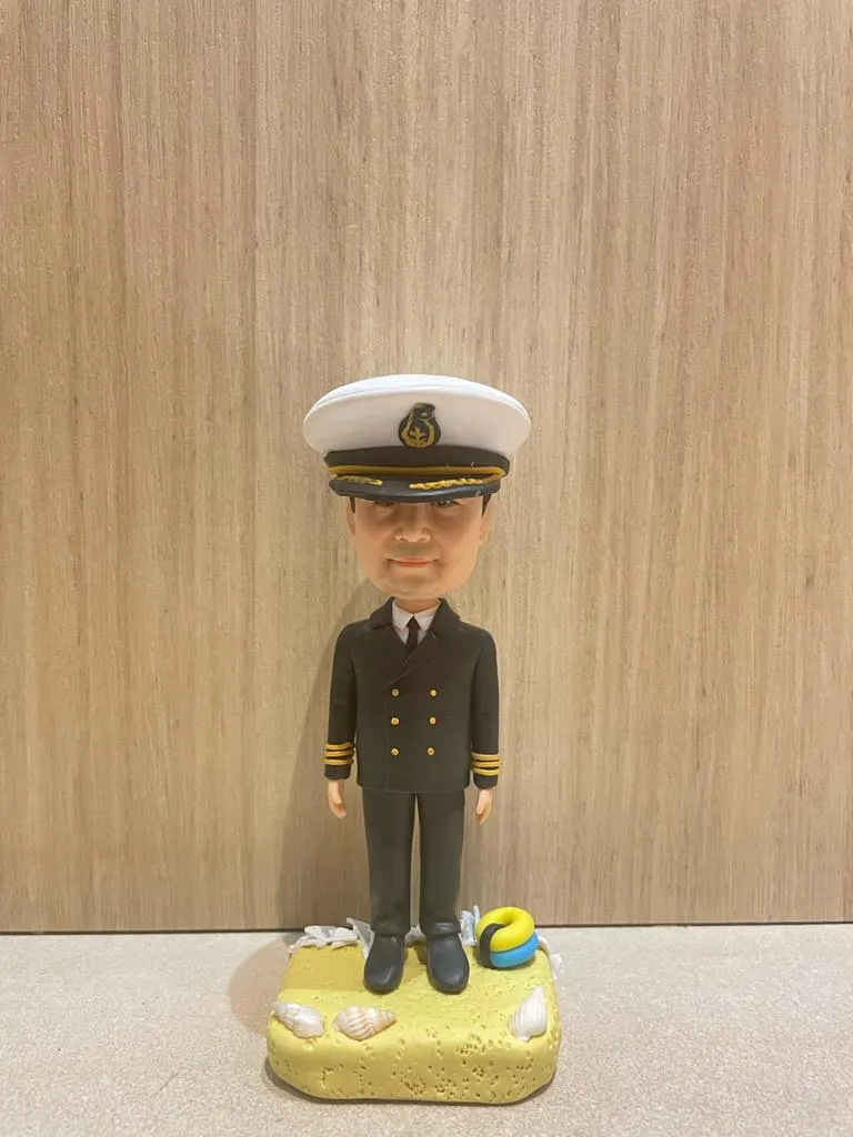 船長主題的人像公仔直立於沙灘質感底座上；戴白色船長帽（黑色帽沿與金色繡飾、帽徽），穿深色雙排扣制服外套與長褲、黑鞋，袖口配金色階級條。底座上點綴貝殼與迷你救生圈等海洋元素。