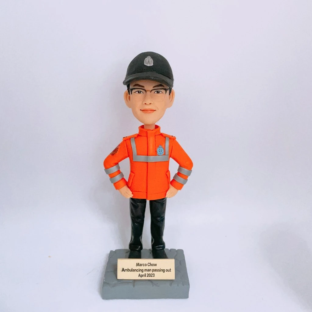 救護主題的人像公仔站在灰色石紋方形底座上，雙手叉腰；公仔戴黑色帽、配眼鏡，穿亮橘色高可視外套，衣身與袖口有銀色反光條與臂章徽章，下著黑色長褲與黑鞋。底座前有金色小牌，刻有英文姓名與「Ambulancing man passing out April 2023」字樣。