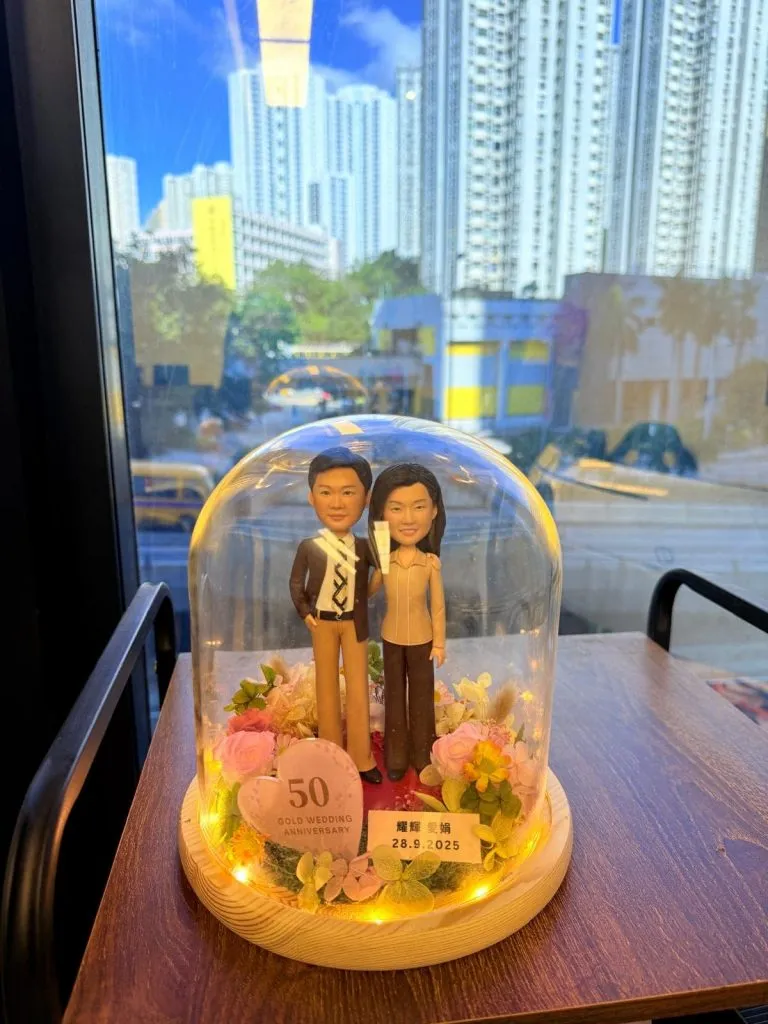一組長輩主題的人像公仔置於玻璃盅內展示:木質圓形底座上鋪紅色地面與綠植苔面,周圍環繞粉、奶白與淡黃系的玫瑰與葉材。兩位角色並肩站立:左側穿深色外套、白襯衫與棕色長褲,右側穿米色上衣與深色褲,皆著深色鞋。玻璃盅內側繞有暖白燈串,底座前方有粉色愛心立牌寫「50 GOLD WEDDING ANNIVERSARY」,旁邊金色小牌標示日期「28.9.2025」與中文字樣。作品置於木桌上,背後可見窗外高樓城市景與車流。