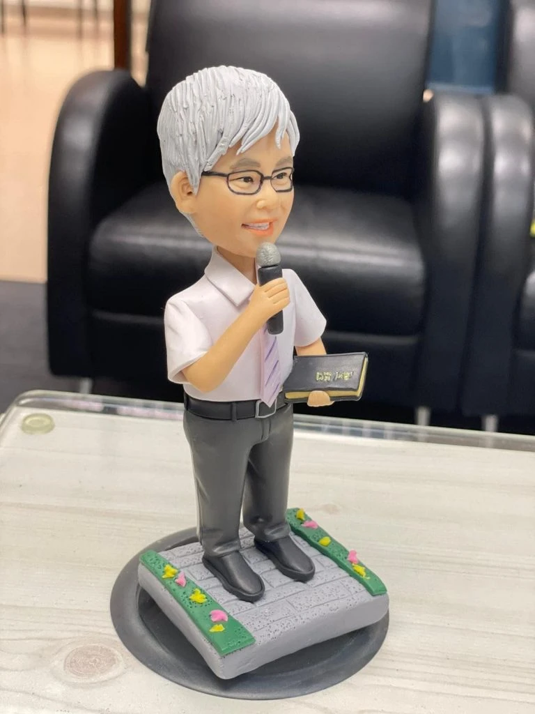 宗教主題的人像公仔：銀灰短髮、戴眼鏡的男性站姿，穿白襯衫配淡紫色領帶與深灰長褲，右手握麥克風，左手持黑色金邊聖經樣小冊；腳踏石磚紋台座，四周綠邊點綴小花。適合作為牧師禮物的人像公仔。