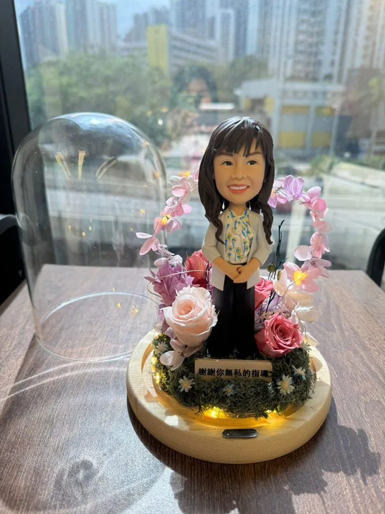 教師主題人像公仔置於木質圓形底座中央，底座內圈繞有暖黃燈串；公仔穿米色外套與碎花襯衫、長髮微卷微笑站立。四周以粉紅與玫瑰色不凋花、苔蘚與小白花環繞，後方形成粉色花環拱門；前方金色小牌寫有「謝謝你無私的指導」。左側放著透明玻璃罩，背景是靠窗的城市景致與校園般建築。