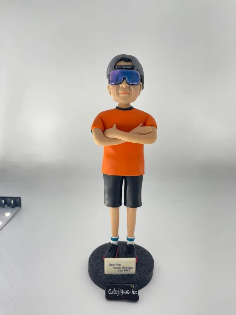 休閒風格的人像公仔雙臂交叉站立於黑色圓形底座上，戴反戴灰色帽、配大型反光墨鏡，穿橘色短袖上衣、深色及膝短褲、藍白襪與黑紅運動鞋；底座前貼有小牌寫著「Dear Wa Happy Birthday Sep 2021」。