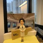 四大會計師所主題的人像公仔：短髮女性穿米色套裝外套與黑色內搭，雙手於身前交握，站在帶有 EY安永 標誌的接待台前；背板呈現現代化辦公室與天花吊燈設計，台前左側有英文字樣的榮休感謝銘牌。場景放置在窗景前的桌面上，整體營造出專業、親切的事務所接待區氛圍，適合作為 EY安永 高層榮休禮物與會計師人像公仔展示。