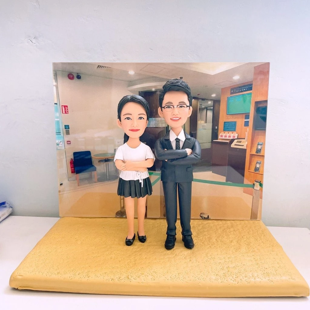 一對成對的人像公仔站在金色質感底座上，背後是銀行或商務大廳風格的背景板。左側公仔為短袖白襯衫搭配深色短裙與黑色便鞋，雙手交叉；右側公仔穿深灰色西裝、白襯衫與領帶，戴眼鏡，雙臂抱胸。場景包含等候區椅子、自助服務機與資訊螢幕。