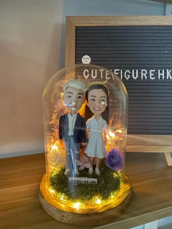 木質底座上的玻璃永生花燈罩作品：內含一對情侶人像公仔，左側角色著深藍外套與破洞牛仔褲、白鞋；右側角色穿白色V領短袖連身裙與白鞋。腳下覆苔綠草，周圍點綴紫玫瑰與乾燥花材，環繞暖白色小燈串；前方小牌寫有日期「2021-11-11」。背景為木桌與置有字母板的場景。