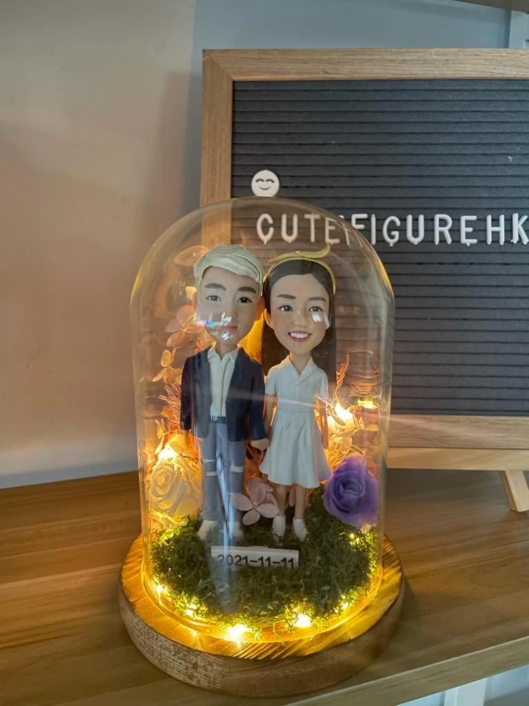 木質底座上的玻璃永生花燈罩作品：內含一對情侶人像公仔，左側角色著深藍外套與破洞牛仔褲、白鞋；右側角色穿白色V領短袖連身裙與白鞋。腳下覆苔綠草，周圍點綴紫玫瑰與乾燥花材，環繞暖白色小燈串；前方小牌寫有日期「2021-11-11」。背景為木桌與置有字母板的場景。