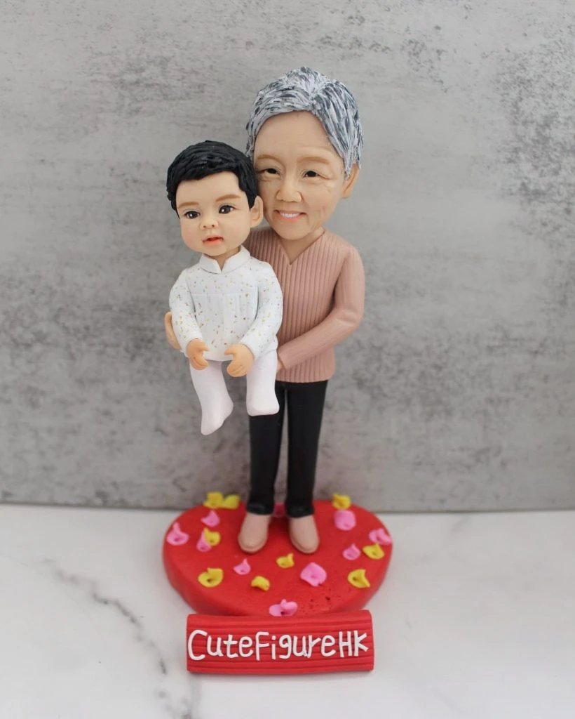 水泥灰背景前，一組祖孫主題人像公仔立於紅色心形底座：年長角色穿粉棕色針織上衣與黑褲、米色鞋，面帶微笑從後方雙手托起懷中的bb；小朋友穿白色點點襯衫與白色褲襪，短髮、神情可愛。底座撒有粉、黃小花瓣裝飾，前方紅色小牌寫「CuteFigureHK」。