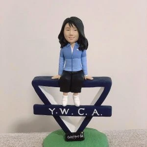 女同事主題的人像公仔坐在深藍色「Y.W.C.A.」字樣的三角造型標誌上；公仔長髮、微笑，穿淺藍外套與深色半身裙、白色鞋子。標誌立於綠色草地質感圓形底座上，前方有小黑牌寫著「cutefigure-hk」。