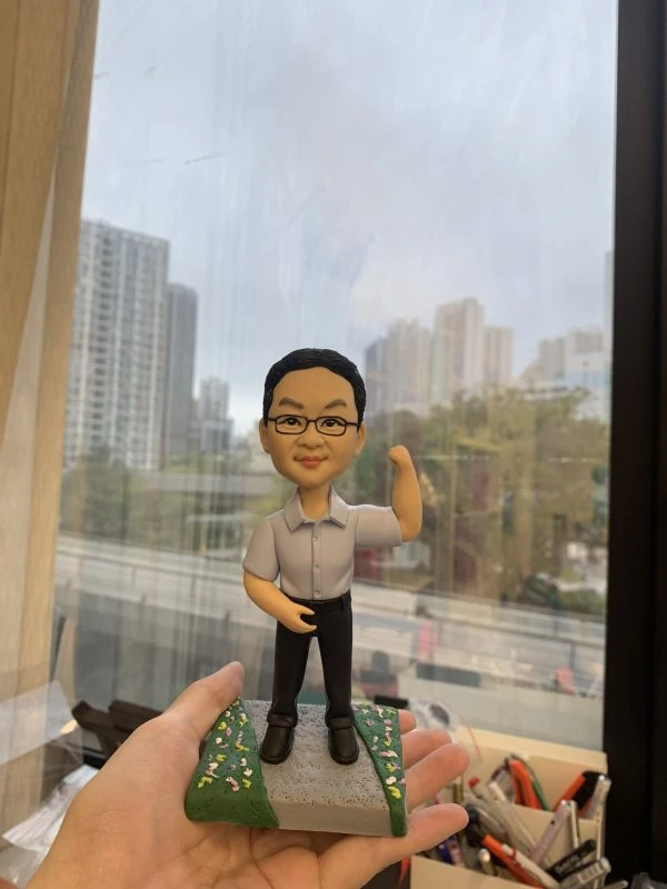 老闆主題的人像公仔立於灰色步道與綠地花卉點綴的底座上，由手掌托起展示。公仔為短髮、配戴眼鏡，穿淺灰短袖襯衫、黑色長褲與黑色皮鞋；右臂上舉作打氣姿勢，左手自然放置。背景為臨窗的辦公環境，可見城市高樓與道路景觀，前景擺有文具收納盒。