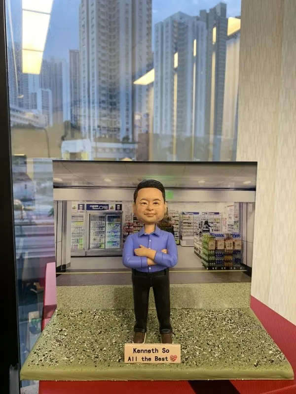 一款老闆主題的人像公仔，站在綠灰色砂石質感底座上，雙臂交叉、藍色長袖襯衫、黑色長褲與棕色鞋。底座前方名牌印有「Kenneth So / All the Best」與表情符號。背板是便利商店室內場景，包含飲料冷藏櫃、貨架與照明；整體置於臨窗環境，窗外可見高樓城市景觀。