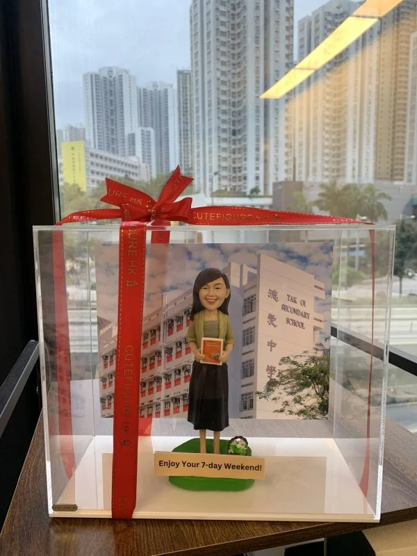 一款放於透明展示盒內並以紅色緞帶蝴蝶結包裝的退休公仔人像公仔。公仔留中長髮，穿淺綠外套、條紋上衣與深色長裙，雙手抱著一本橘色書本；腳邊有小型花束造型，站在綠色草地底座上。背景板為白色與紅色相間的中學校舍立面，右側可見「TAK OI SECONDARY SCHOOL」及中文校名。前方牌匾寫有「Enjoy Your 7-day Weekend!」。整體置於木紋桌面，窗外是高樓城市景觀。