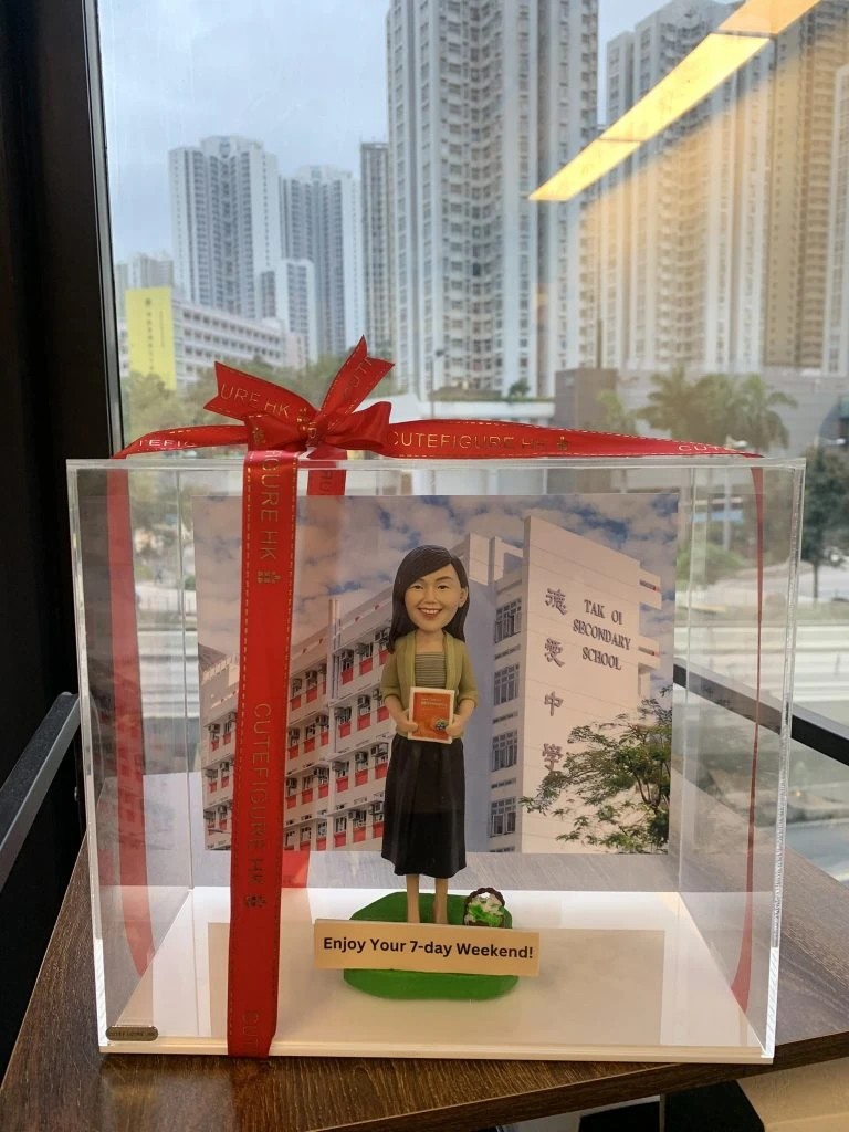 一款放於透明展示盒內並以紅色緞帶蝴蝶結包裝的退休公仔人像公仔。公仔留中長髮,穿淺綠外套、條紋上衣與深色長裙,雙手抱著一本橘色書本;腳邊有小型花束造型,站在綠色草地底座上。背景板為白色與紅色相間的中學校舍立面,右側可見「TAK OI SECONDARY SCHOOL」及中文校名。前方牌匾寫有「Enjoy Your 7-day Weekend!」。整體置於木紋桌面,窗外是高樓城市景觀。