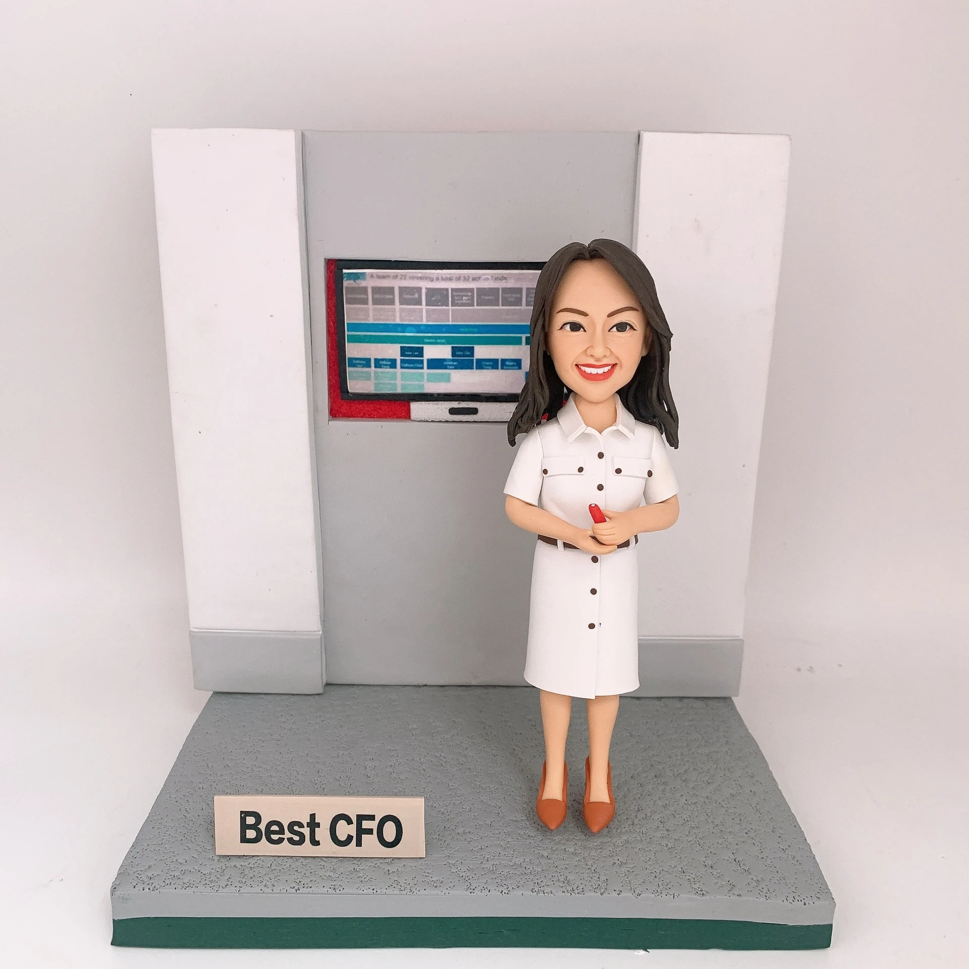 灰白色辦公背板前的CFO主題人像公仔：長深色髮女性公仔穿白色襯衫式連身裙，腳踩棕色尖頭鞋，雙手握紅色筆微笑站立。身後嵌入式螢幕顯示藍灰色表格與儀表板介面。公仔立於灰色紋理底座，前方放置金屬色小名牌寫有「Best CFO」。背景為純白。