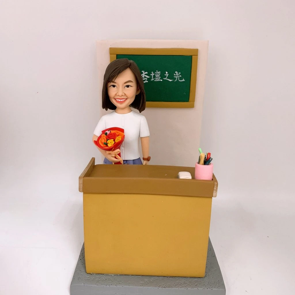 教室場景的人像公仔：一位短髮女性公仔站在黃褐色講台後方，右手捧著橙紅色花束，左側講台上放著粉筆，右側是一個粉色筆筒插著彩色筆。背後掛著綠色小黑板，上寫白字「杏壇之光」。整體置於灰色階梯式底座，背景為素白牆。