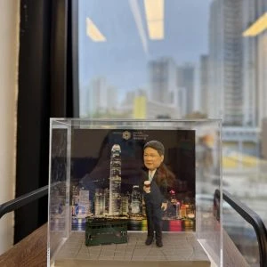 一款人像公仔置於透明展示盒中，背景為維多利亞港夜景與高樓天際線；地台為灰色石板路紋理。公仔穿深色西裝、白襯衫與條紋領帶，右手拿外帶咖啡杯，站姿輕鬆。腳邊擺放一台深綠色雙層電車模型；背景板可見英文字樣與標誌。整體放在木層板上，窗外是城市高樓景致。