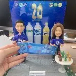 飲品品牌20週年主題的人像公仔場景：一男一女公仔穿著藍色K2品牌T恤與球鞋，微笑站立，雙手共同舉著藍色宣傳橫幅（印有K2與NO.1字樣）；右側白色圓桌擺放多款彩色瓶裝飲品模型。背板是藍底「20th LIFE WATER 20th ANNIVERSARY」設計，並有多款瓶裝水與飲料圖像；地台為灰色地磚，前方小牌寫有“Congratulations! From: Hong Kong Cousin (2023.8.13)”。整體呈現慶祝品牌里程碑的紀念公仔擺設。