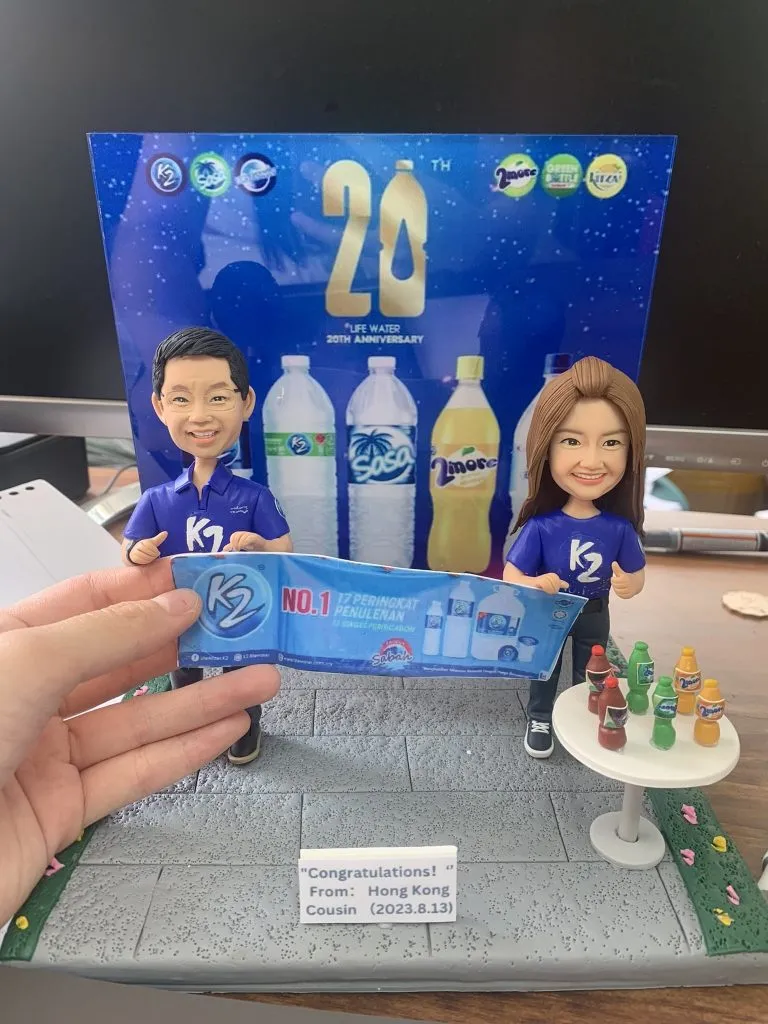 飲品品牌20週年主題的人像公仔場景：一男一女公仔穿著藍色K2品牌T恤與球鞋，微笑站立，雙手共同舉著藍色宣傳橫幅（印有K2與NO.1字樣）；右側白色圓桌擺放多款彩色瓶裝飲品模型。背板是藍底「20th LIFE WATER 20th ANNIVERSARY」設計，並有多款瓶裝水與飲料圖像；地台為灰色地磚，前方小牌寫有“Congratulations! From: Hong Kong Cousin (2023.8.13)”。整體呈現慶祝品牌里程碑的紀念公仔擺設。