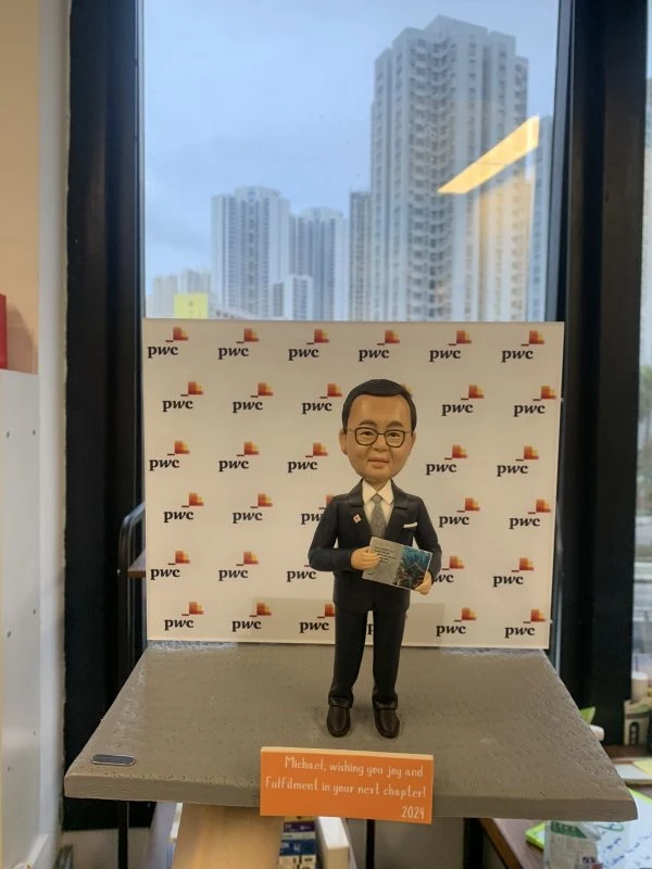 人像公仔站在灰色台座上，身穿深色西裝與領帶，配戴眼鏡，手持一本印有城市景觀圖的冊子；背板重複出現「pwc」與彩色方塊標誌，前緣橘色小牌寫有祝福語與年份2024；場景位於室內窗前，可見遠處高樓天際線。整體呈現專業商務與顧問氛圍的人像公仔展示。