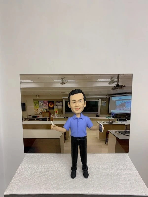 教室主題的人像公仔：短髮男性公仔身穿藍色短袖襯衫與黑色長褲，雙臂微張站立，右手持白色粉筆、左手拿書本。公仔置於顆粒感白色台座前，背板為教室場景，包含黑板、投影幕、講桌、實驗桌與牆上海報。