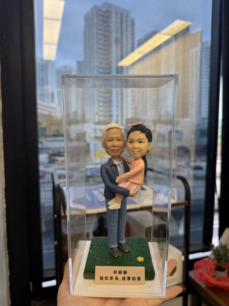 透明展示盒內的一組人像公仔：一位長者造型穿藍色外套、襯衫與長褲，雙手抱著一位穿粉色洋裝與髮飾的小朋友／bb公仔；兩人站在草地質感的綠色方形底座上，底座前有白色名牌印有祝福字樣。背景為室內窗邊，可見高樓與室內陳設的模糊景象。