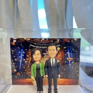 兩款並立的人像公仔置於白色砂岩質感底座上，背板為舞台與金色彩帶燈光效果。左側女性公仔留短捲髮，穿綠色外套配黑色上衣與長褲、灰色鞋，右手持米色手袋；右側男性公仔戴眼鏡、穿黑色西裝與白襯衫、黑黃條紋領帶，胸前別花。前方金色名牌刻有中文祝賀字樣，內容包含「祝賀××校監…榮休愉快 2024年7月」等。整體呈現典禮與賀壽氣氛。